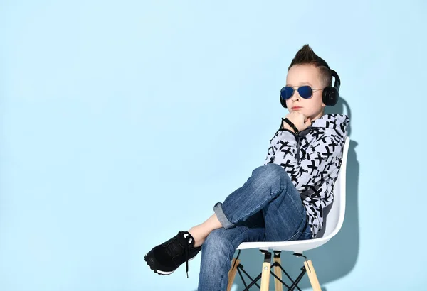 Tonårsbarn i hörlurar, solglasögon, blå jeans, luvtröja, sneakers. Stöter hakan i knytnäven, sitter på stolen. Blå bakgrund — Stockfoto