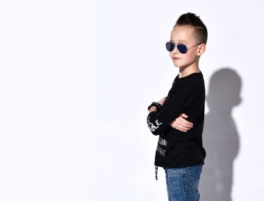 Güneş gözlüklü genç ve siyah kazaklı. Üzerinde yazılar, kot pantolon ve bilezik var. Beyaz stüdyo arka planında yalnız poz vermiş.