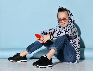 Güneş gözlüklü çocuk, kot pantolon ve kapüşonlu, siyah bilezik, spor ayakkabı. Kırmızı akıllı telefon kullanarak, beyaz zeminde oturarak. Mavi arkaplan