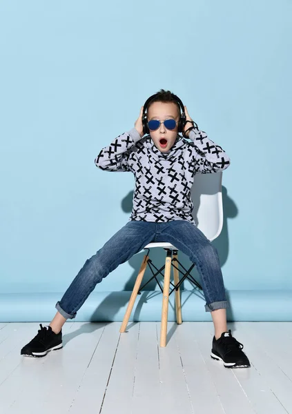 Adolescente con auriculares, gafas de sol, ropa casual. Mirando maravillado, escuchando música. Sentado en una silla blanca. Fondo azul — Foto de Stock