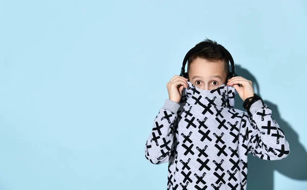 Niño adolescente con auriculares y brazalete negro. Él está escondiendo su boca con sudadera con capucha elegante mientras posa sobre fondo azul —  Fotos de Stock