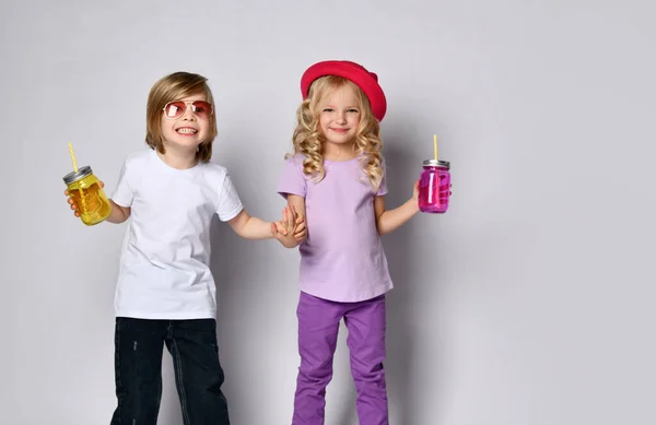 Маленька дівчинка і хлопчик в барвистому повсякденному одязі. Усміхаючись, тримаючи руки, жовті та рожеві пляшки коктейлів. Позування ізольовано на білому — стокове фото