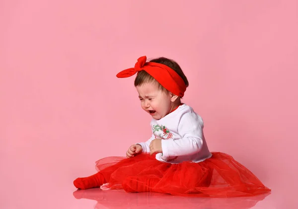 Toddler in white bodysuit, red headband, poofy skirt. She crying, sitting on floor against pink studio background. Close up — Stock Photo, Image
