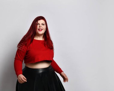 Kırmızı kazıklı, siyah sutyen ve deri etekli obez kadın. Dans ediyor, beyaz arka planda yalnız poz veriyor.
