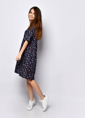 Siyah elbiseli, çiçekli, beyaz ayakkabılı esmer bir manken. Gülümsüyor, yüzüne dokunuyor, beyazlar içinde yalnız poz veriyor. Tam uzunluk