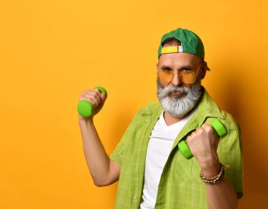 Büyükbaba yeşil şapkalı ve gömlekli, beyaz tişörtlü, bilezik takıyor. Gülümsüyor, halterlerle egzersiz yapıyor, turuncu arka planda poz veriyor.