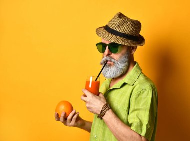 Şapkalı, yeşil gömlekli, güneş gözlüklü yaşlı bir adam. Turuncu arka planda durarak, portakal suyu tutarak ve tüple taze meyve suyu içerek.