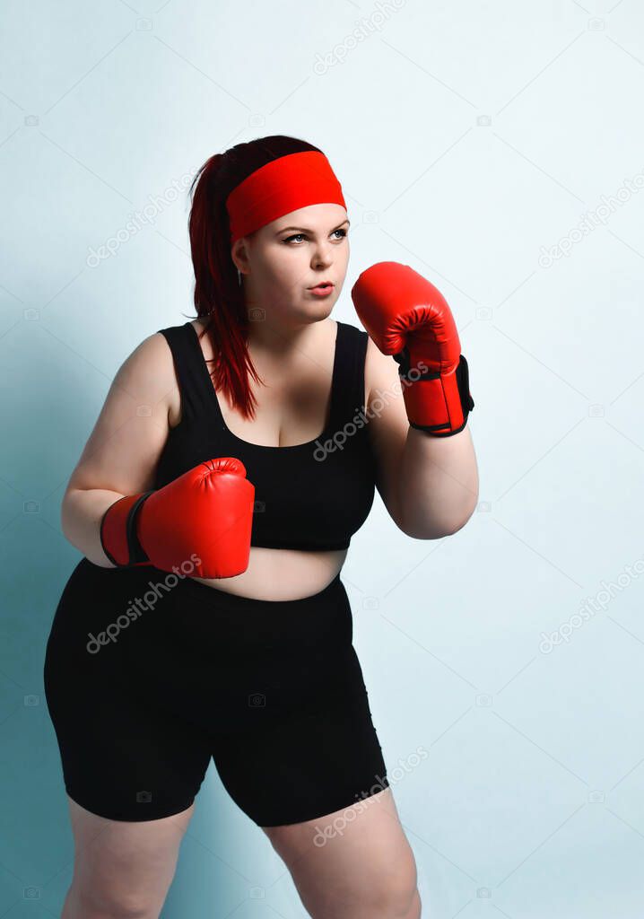 Thoughtful plump young girl in black sporty top, shorts and red boxing gloves looks up thinking over strategy in forthcoming fight. Copy space