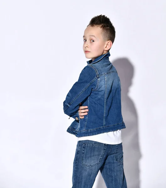Teenage dítě v tričku, modré džíny, džínová bunda. Stojí zády ke kameře, ohlíží se, pózuje izolovaně na bílém — Stock fotografie