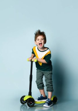 Renkli tişörtlü, gri kotlu ve spor ayakkabılı küçük esmer manken. Mavi arka planda, scooter 'ın üzerinde dikilirken çığlık atıyor.