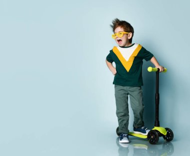 Renkli tişört giyen neşeli çocuk, sarı gözlük, gri kot pantolon, spor ayakkabı. Adam motosikletinin yanında duruyor.