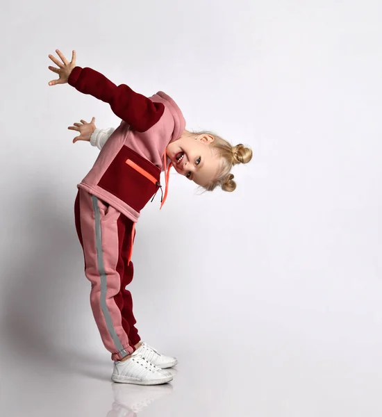 Lachende blonde schoolmeisje in roze en rode jympak leunend hoofd naar voren nemen van de armen terug. Full length portret geïsoleerd op lichtgrijs — Stockfoto