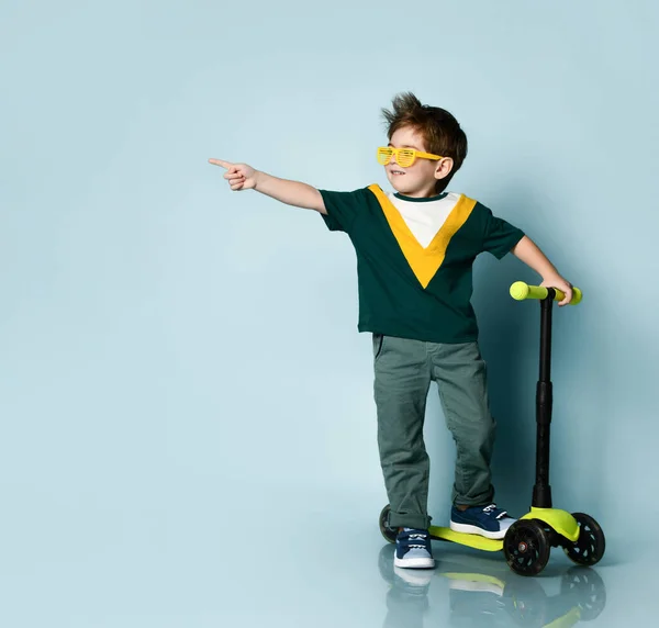 Chlapec v barevném tričku, žlutých brýlích, džínách, teniskách. S křikem zvednutou rukou, stojící na koloběžce. Modré pozadí — Stock fotografie