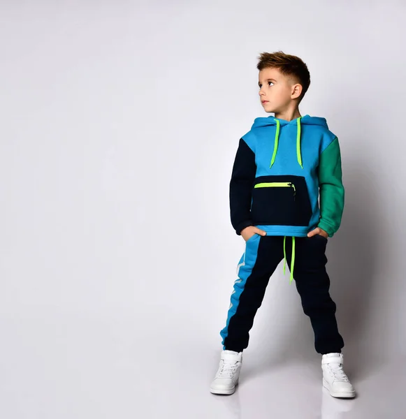 Kind in buntem Trainingsanzug, Turnschuhen. Er steckte die Hände in Taschen und posierte isoliert auf weißem Studiohintergrund. Mode — Stockfoto