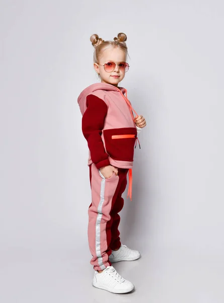 Klein blond model met kroonkapsel, in zonnebril en een kleurrijk trainingspak. Ze poseert in volle groei poserend geïsoleerd op wit.. — Stockfoto