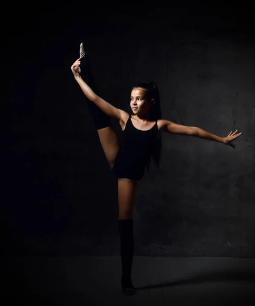 Giovane ginnasta sorridente ragazza in corpo nero sport e calzature speciali in piedi, tenendo la gamba tesa in mano e mantenendo l'equilibrio — Foto Stock