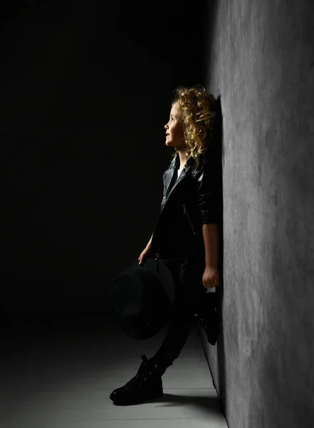 Lächelndes kleines Mädchen mit blonden Locken im schwarzen Lederanzug, das im Scheinwerferlicht nahe der dunklen Wand steht, nach oben blickt und den Hut in der Hand hält — Stockfoto