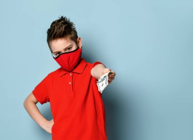 Parlak kırmızı tişörtlü ve tıbbi maskeli ciddi bir çocuk elinde kalça ile cesurca poz veriyor, kameraya para teklif ediyor, mavi üzerine izole edilmiş.