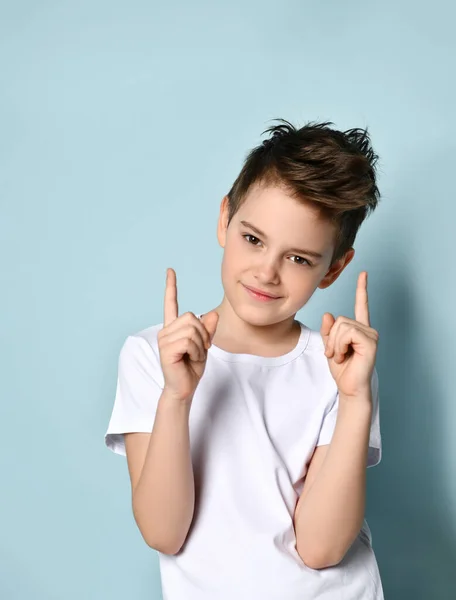 Agradable amigable niño en camiseta blanca mirando hacia adelante con sonrisa poniendo ambos dedos índice hacia arriba. Retrato de media longitud aislado en azul claro —  Fotos de Stock