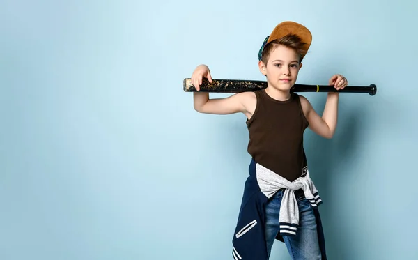 Adolescente con gorra, camiseta, jeans y sudadera con capucha atados alrededor de su cintura. Sostiene bate de béisbol negro, posando sobre fondo azul —  Fotos de Stock