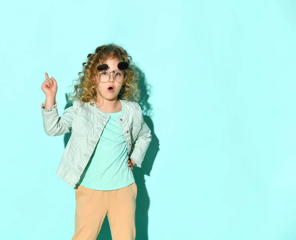 Niño pequeño con gafas de sol, chaqueta vaquera y pantalones beige. Levantó su dedo índice, posando sobre fondo turquesa — Foto de Stock