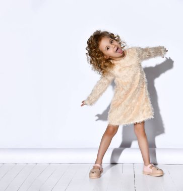 Pembe, kıvırcık, sarı elbiseli ve sandaletli neşeli kız kollarını ve bacaklarını açarak şakalaşıyor ve uzanıyor. Beyaz üzerine izole edilmiş tam boy portre