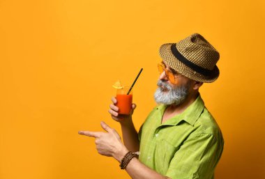 Büyükbaba şapkalı, yeşil gömlekli, güneş gözlüklü. Bıyık bükme, elinde taze sıkılmış meyve suyu bardağı, turuncu arka planda poz verme.