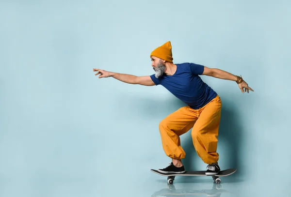 Aged male in t-shirt, orange pants, hat, gumshoes. Riding black skateboard, clenched fists, posing sideways on blue background