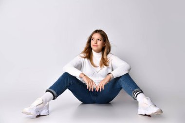 Kot pantolon, kazak, çorap ve spor ayakkabı giyen sarışın ergen. Gülümsüyor, yerde oturuyor, bacakları açık, bembeyaz. Kapat.