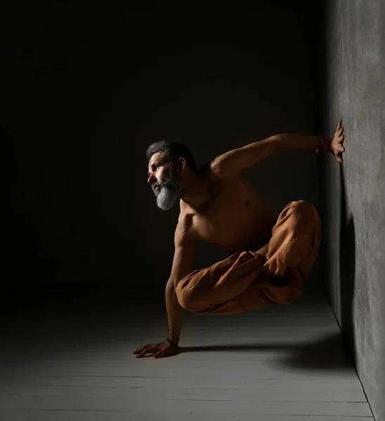 Hombre viejo, torso desnudo, en pantalones marrones está practicando yoga. Piernas en pose de loto, colocando una mano contra la pared, otra en el suelo — Foto de Stock