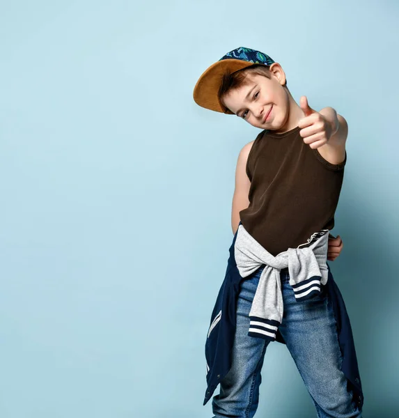 Teenager v čepici, hnědé tričko, džíny a mikina kolem pasu. Usmívat se, ukazovat palec nahoru, pózovat na modrém pozadí — Stock fotografie