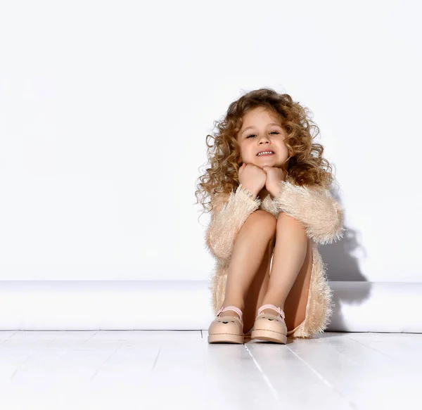 Adorabile bambina con i capelli ricci dorati seduta sul pavimento con sorriso sognante appoggiando il mento con le mani. Close up ritratto isolato su bianco — Foto Stock
