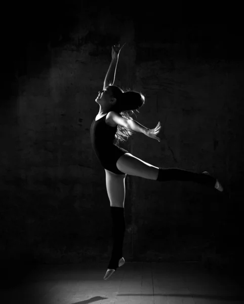 Young girl gymnast in black sport body and special uppers jumping in gymnastic pose over dark background — 스톡 사진