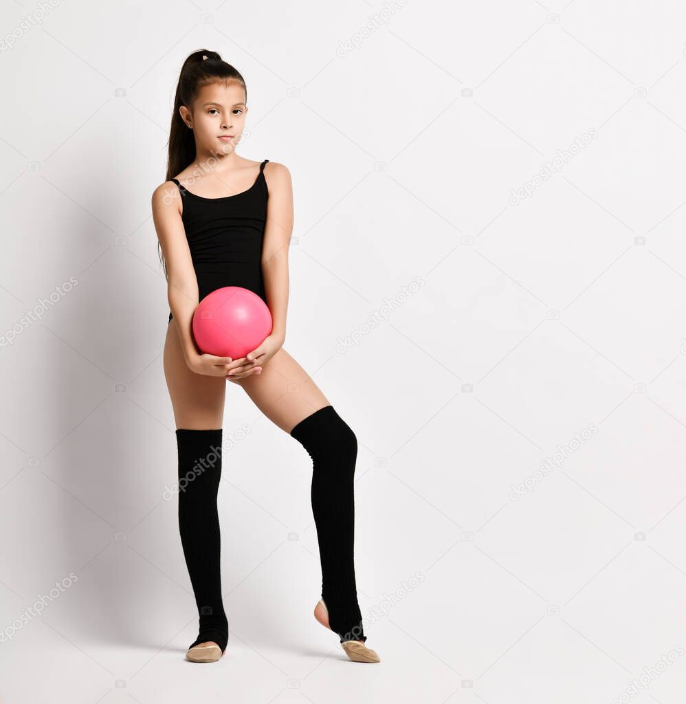 Young girl gymnast with long hair in black sport body and uppers standing and holding pink gymnastic ball in hands