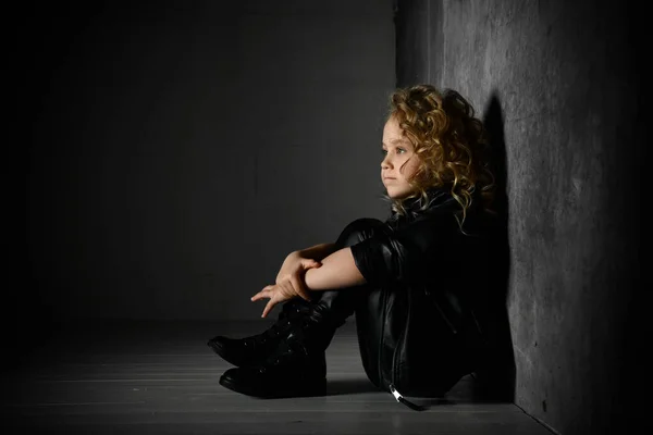 Menina adorável com cabelo encaracolado dourado vestido de preto sentado em destaque no chão perto da parede escura, sorrindo calmamente segurando a mão no joelho . — Fotografia de Stock