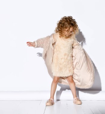 Pembe elbiseli, kıvırcık saçlı ve sandaletli sevimli sarışın kız şık tüylü bir ceket giyiyor. Beyaz üzerine izole edilmiş tam boy portre