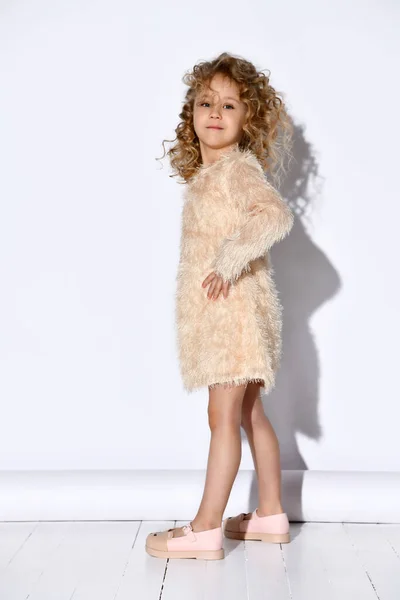 Little blonde curly kid in fluffy pink dress and shoes. She has put her hands on hips, posing standing sideways isolated on white