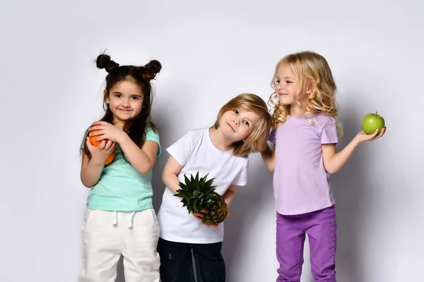 Chłopiec i dziewczynki w kolorowych, luźnych ciuchach. Śmiech, trzymanie ananasa, pomarańczy i zielonych jabłek. Pozowanie na białym — Zdjęcie stockowe
