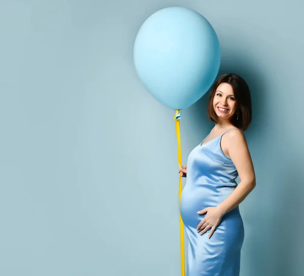 파란 드레스를 입은 임신 한 여성. 웃고, 배에 손을 얹고, 노란 리본으로 풍선을 들고, 파란 배경에 옆으로 눕고 — 스톡 사진