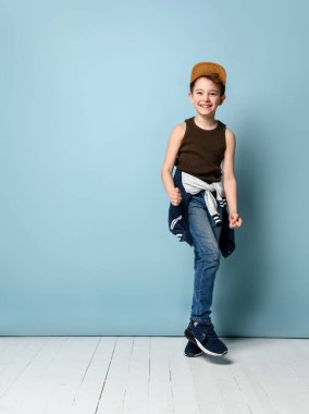 Şapkalı, kahverengi atletli, kot pantolonlu, beline ve spor ayakkabılarına kapüşonlu bir genç. Gülümsüyor, mavi arka planda poz veriyor