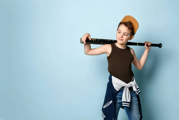 Adolescente con gorra, camiseta, jeans y sudadera con capucha atados alrededor de su cintura. Sostiene bate de béisbol negro, posando sobre fondo azul — Foto de Stock