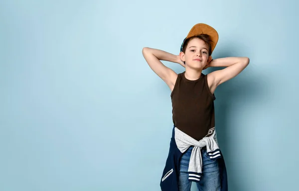 Niño activo vestido con estilo deportivo se para mirando a un lado y tocando la tapa de su cabeza, creando una broma o travesura. Estudio tiro aislado en azul —  Fotos de Stock