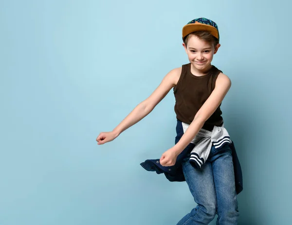 Tonårspojke i mössa, brun undertröja, jeans och huvtröja runt midjan. Le och dansa medan poserar på blå bakgrund — Stockfoto