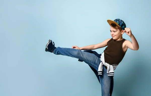 Muchacho adolescente brutal en jeans, camisa y gorra sin mangas haciendo patada lateral y balanceando un puñetazo listo para luchar. Retrato aislado sobre fondo azul — Foto de Stock