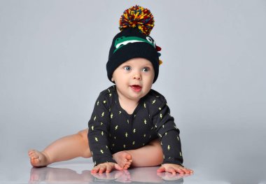 Siyah elbiseli, şapkalı, timsah izli, yalınayak bir çocuk. Gülümsüyor, beyaz zeminde tek başına oturuyor. Kapat.