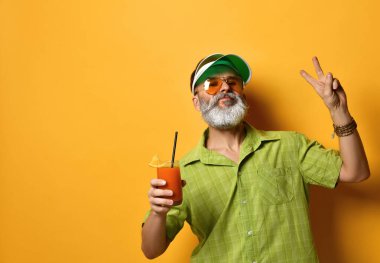 Yeşil güneşlik, gömlek ve güneş gözlüklü sakallı adam. Bir bardak sıkılmış meyve suyu, sıkılmış yumruk, turuncu arka planda poz.