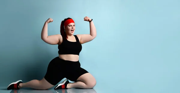 Plus size rothaarige Frau in rotem Stirnband, schwarzem Oberteil, kurzer Hose, Turnschuhen. Muskeln zeigen, auf Knien vor blauem Hintergrund stehen — Stockfoto