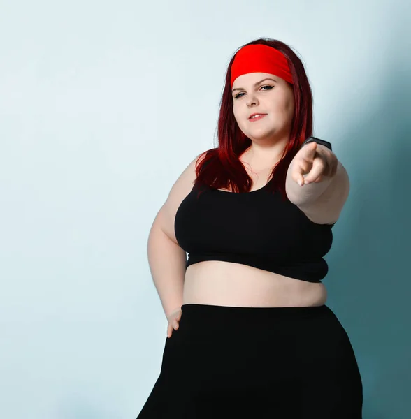 Overweight ginger girl in red headband, black top, leggings, sneakers.  Showing stop sign by her palm, posing on blue background Stock Photo