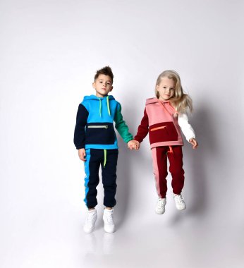 Çocuklar, erkek ve kız kardeşler, renkli eşofmanlar ve spor ayakkabılar içinde. El ele tutuşuyorlar, zıplıyorlar, beyaz arka planda soyutlanıyorlar.