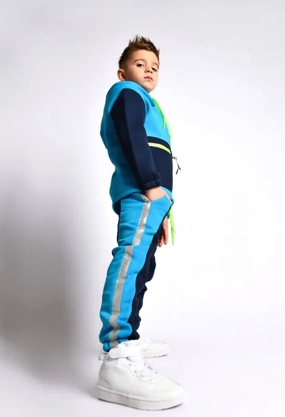 Side view shot of little schoolboy in bright cotton tracksuit standing with hands in pockets looking down. Full length portrait isolated on light grey — Stock Photo, Image